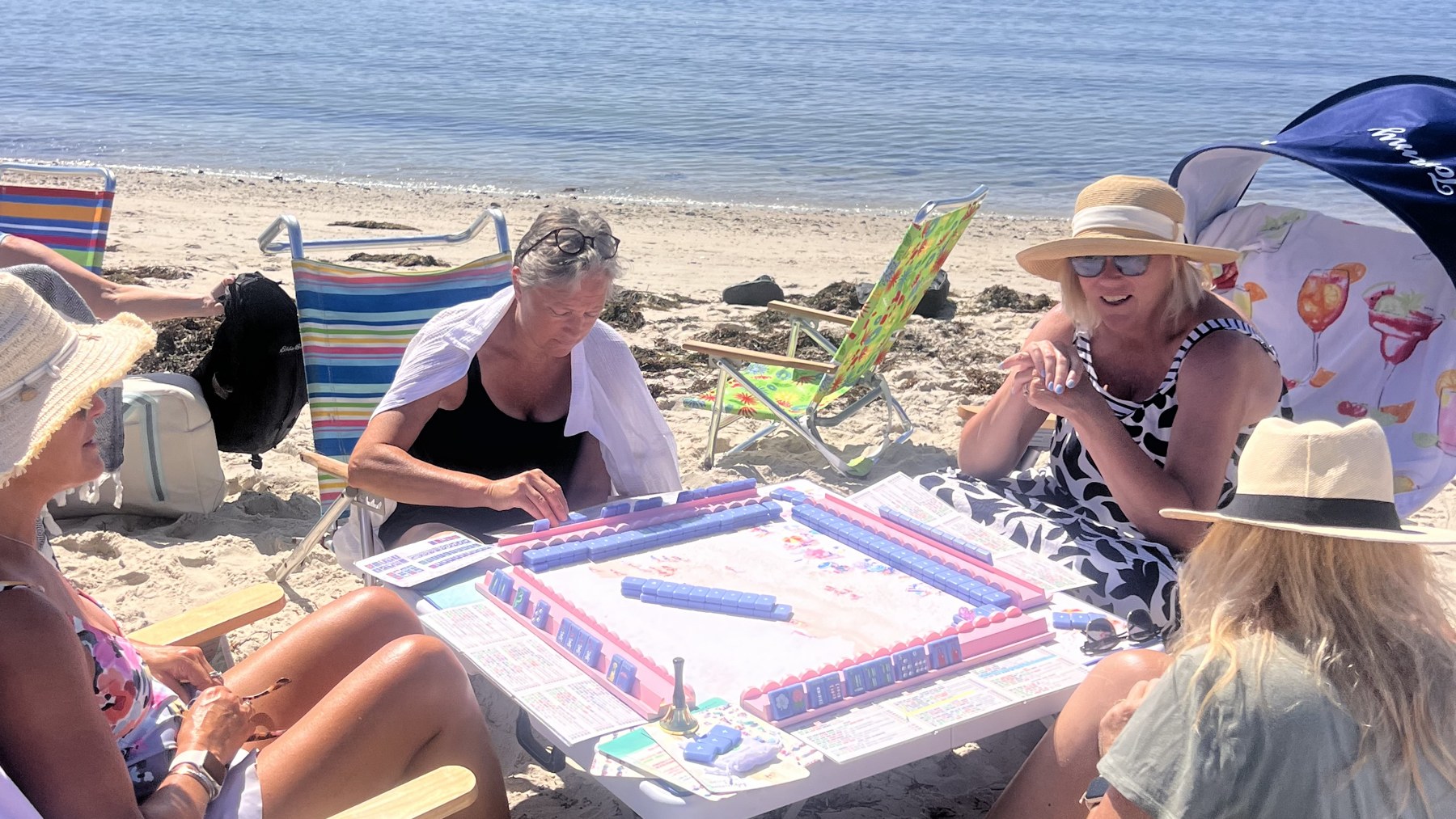 Four friends playing American mahjong around a table on the sand at the beach, with the ocean behind them